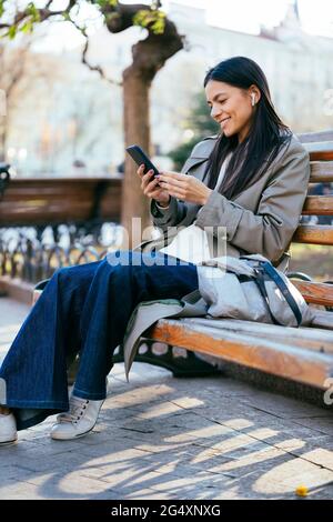 Lächelnde Frau, die im öffentlichen Park auf der Bank sitzt und einen Mantel mit einem Smartphone trägt Stockfoto