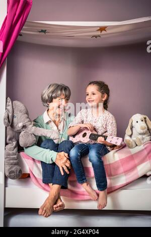 Enkelin Gitarre spielen während der Sitzung von Großmutter auf dem Bett Stockfoto