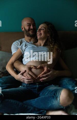 Glücklicher Mann macht Herz Hand Zeichen auf Bauch der Schwangeren zu Hause Stockfoto