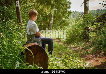 Ein europäischer Teenager in einem weißen T-Shirt sitzt auf einem Baumstamm im Wald Stockfoto