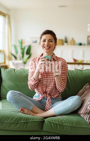 Glückliche junge Frau, die zu Hause auf einer Couch sitzt. Die Frau trinkt Tee. Stockfoto