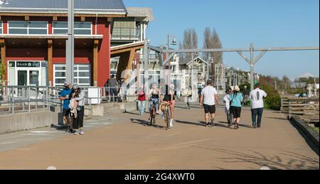 Straßenansicht der Menschen mit Gesichtsmasken, die auf dem Bürgersteig am Flussufer in Guilford, BC, Kanada, spazieren und Fahrrad fahren, April 18,2021. Stockfoto