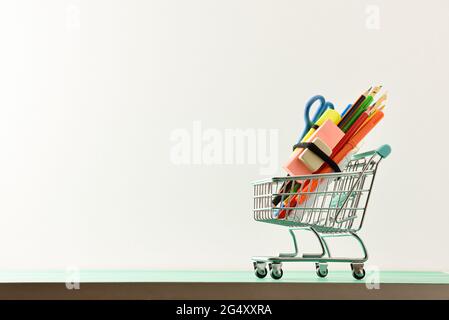 Schulbedarf-Shopping-Konzept mit Mini-Warenkorb voller Schulbedarf auf grünem Tisch mit weißem isolierten Hintergrund. Vorderansicht. Stockfoto