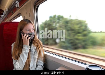 Unterwegs weibliche Teenager in Schutzmaske sprechen auf Handy, während in der Nähe Fenster im Zug sitzen während der Sommerreise durch die Landschaft A Stockfoto