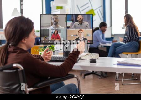 Behinderter gelähmter, ungültiger Mitarbeiter während eines virtuellen Meetings, der am Start-up-Büro über eine Webcam mit Partnern online diskutiert. Geschäftsfrau auf Videokonferenz. Stockfoto