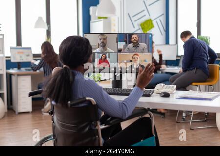 Gelähmte, behinderte schwarze Angestellte, die im Rollstuhl sitzend immobilisiert sitzt und im Geschäftsbüro Online-Videomeeting mit Kollegen aus der Ferne diskutierte. Ungültiger Mitarbeiter, der in der Finanzgesellschaft arbeitet. Stockfoto