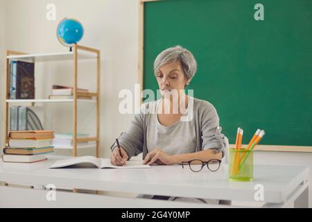 Die Lehrerin schreibt, während sie in der Schule am Schreibtisch im Klassenzimmer mit einem grünen Brett sitzt. Bildungsunterricht. Stockfoto