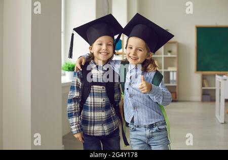 Zwei glückliche Grundschüler in akademischen Kappen lächeln und geben Daumen nach oben Stockfoto