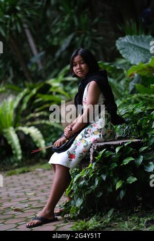 Ein süßes Thai-Mädchen im Teenageralter, das ein Satinkleid mit Fancywork und Duftstäbchen in der Hand trägt, ruht sich in einem tropischen Garten des buddhistischen Tempels aus Stockfoto