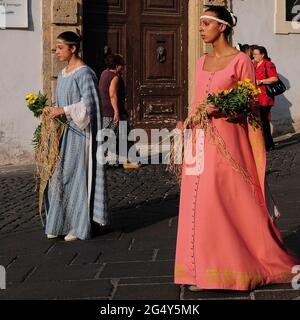 Zwei anmutige junge Damen in eleganten, lachsrosa und hellblauen, langen Kleidern tragen Blumensprays, während sie durch die Piazza Guglielmo Marconi in Agagni, Latium, Italien, spazieren. Der historische Festzug im August ehrt den schutzheiligen der Stadt, den christlichen Bischof und Märtyrer aus dem 3. Jahrhundert n. Chr., den heiligen Magnus von Anagni. Die antike Stadt auf einem Hügel, etwa 71 km (44 Meilen) südöstlich von Rom, war einst die Sommerresidenz römischer Kaiser und mittelalterlicher Päpste. Stockfoto