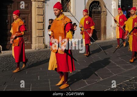 Soldaten in leuchtend roten, orangefarbenen und gelben Kostümen mit Schwertern und Lanzen gehen durch die Piazza Guglielmo Marconi in Anagni, Latium, Italien, und folgen den Schlagzeugknaben des Castello Contrada, einem traditionellen Stadtbezirk, der an einem Festzug und einer Prozession zu Ehren des christlichen Bischofs und Märtyrers aus dem 3. Jahrhundert n. Chr. teilnimmt, Magnus oder Magno von Anagni. Die Männer tragen spitze Kuhhautschuhe, dunkelrote Leggings und rote Stoffkappen und die Schwertkämpfer tragen Lederpanzerung. Die antike Hügelstadt in der Nähe von Rom war einst die Sommerresidenz römischer Kaiser und mittelalterlicher Päpste. Stockfoto