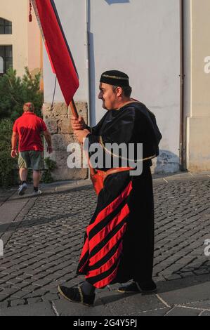 Kostümierte Pracht trifft auf modernen Tourismus als Standardträger mit schwarzem Samt, der mit leuchtend roten Streifen durch die Piazza Guglielmo Marconi in Anagni, Latium, Italien, geht, bei einem Festzug, der an die Vergangenheit der Stadt als Sommerrückzug der römischen Kaiser und Päpste erinnert und ihren Schutzheiligen ehrt, 3. Jahrhundert n. Chr. Bischof und christlicher Märtyrer San Magno di Anagni (Heiliger Magnus von Anagni). Der reich gekleidete Bannerträger trägt das Banner oder die Flagge der Cellere Contrada, eines der traditionellen Bezirke von Anagni. Stockfoto