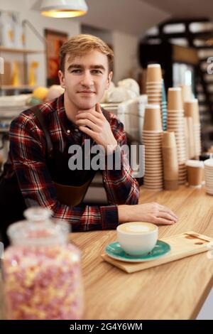 Fröhlicher, männlicher Kellner auf der Schürze, der eine Tasse heißes Getränk hielt Stockfoto