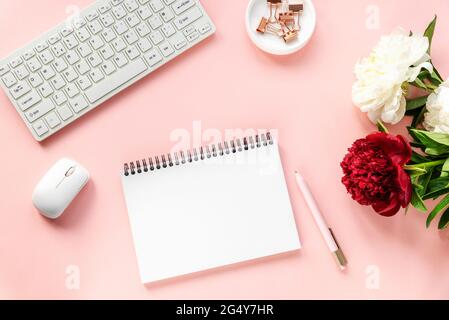 Frauenarbeitsplatz mit Pfingstrosenblumen, leerem Notizblock, Computertastatur, Computermaus und Stift. Draufsicht, flach liegend. Rosa Hintergrund. Stockfoto