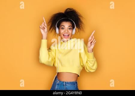 Fröhliches Mädchen in Kopfhörer, hebt die Hand in Tanzbewegung und singt laut auf gelbem Hintergrund Stockfoto