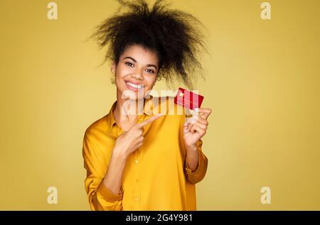 Überfröhliches Mädchen zeigt den Finger auf die Kreditkarte. Foto von afroamerikanischen Mädchen auf gelbem Hintergrund Stockfoto