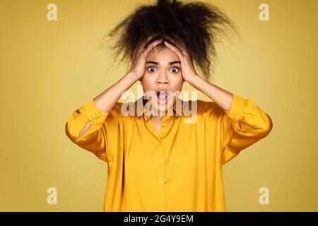 Geschocktes Mädchen hält die Hände auf den Kopf, starrt vor Schrecken. Foto von afroamerikanischen Mädchen auf gelbem Hintergrund Stockfoto