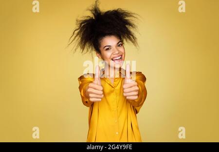 Glückliches Mädchen zeigt Daumen nach oben mit beiden Händen, wie Geste. Foto von afroamerikanischen Mädchen auf gelbem Hintergrund Stockfoto