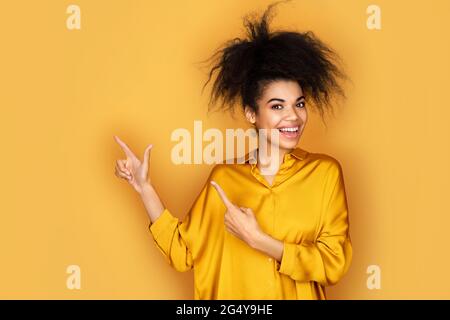 Lächelndes Mädchen zeigt mit den Zeigefingern weg, zeigt auf dem Kopierplatz für Ihren Text. Foto von afroamerikanischen Mädchen auf gelbem Hintergrund Stockfoto