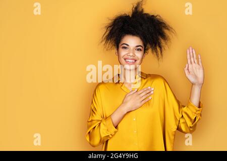 Ehrliches lächelndes Mädchen hält eine Hand am Herzen und hebt die Handfläche, verspricht oder schwört, etwas zu tun. Foto von afroamerikanischen Mädchen auf gelbem Hintergrund Stockfoto