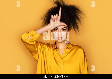 Mädchen, die sich über Menschen mit Fingern auf der Stirn lustig macht, indem sie losere Geste spöttisch und beleidigend macht. Foto von afroamerikanischen Mädchen auf gelbem Hintergrund Stockfoto