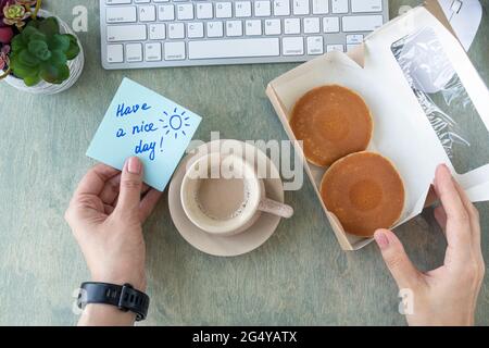 Weibliche Hände halten Notiz mit Worten HABEN EINEN SCHÖNEN TAG, Pfannkuchen in Papierbox, Tasse Kaffee, Tastatur, Kaktus und Mause auf einem Holztisch. Konzept für einen guten Start in den Tag. Draufsicht auf dem Desktop. Flach liegend Stockfoto