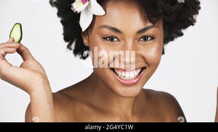 Lächelnde afroamerikanische Frau mit Orchidee im Haar, die Gurkenscheibe auf Grau isoliert hält Stockfoto
