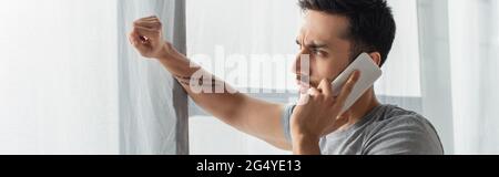 Seitenansicht eines jungen Mannes, der auf dem Smartphone in der Nähe des Fensters sprach, Banner Stockfoto