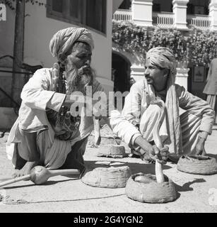 1950s, historisch, zwei Schlangenbeschwörer, die auf dem Boden hocken, einer hält den Hals seiner Schlange, eine Kobra, deren Kopf aus einem kleinen Korb, Indien, hervortritt. Der Charmer oder Heiler mit einem langen Bart hatte eine Pythonschlange um seinen Hals und sein Pongi, ein Musikinstrument, mit dem die Kobra hypnotisiert wurde, sitzt auf dem Boden. Stockfoto