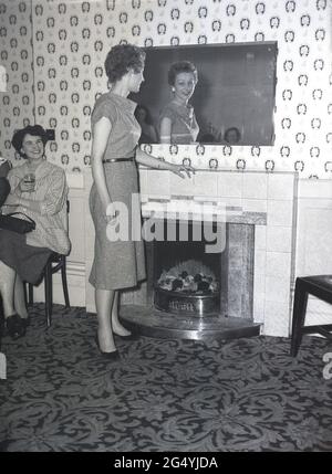 1957, historisch, bei einer Party im Victoria Hotel, Leeds, eine junge Dame in einem eleganten Kleid, die am Kamin steht und in einen Spiegel blickt. Stockfoto