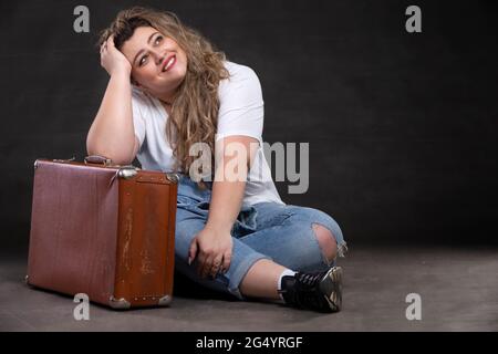 Schöne fette Frau in Denim-Kleidung mit einem alten Retro-Koffer auf grauem Hintergrund. Plus Größe Modell Mädchen. Stockfoto