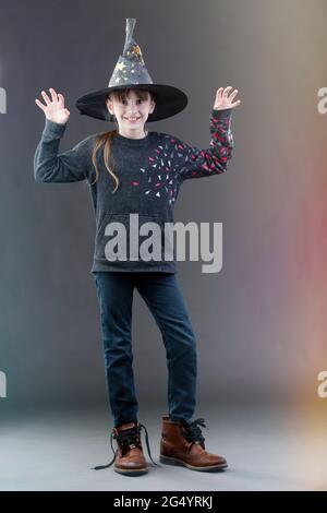 Kleines Mädchen in einem Hexenhut auf grauem Hintergrund. Das Kind auf dem heiligen Fest. Stockfoto