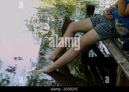 Männliche Füße im Wasser in einem überfluteten Park, Nahaufnahme. Ein junger Mann sitzt auf einer Bank, seine Füße liegen im Wasser und hält ein Telefon in den Händen. Stockfoto