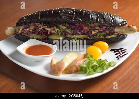 Langer schwarzer Hot Dog mit gebratener Hühnerwurst mit Salat, auf einem Teller mit Sauce, Tomaten und Kartoffeln Stockfoto