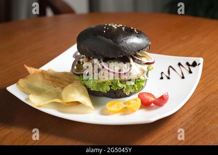 Großer schwarzer Burger mit gegrilltem Fleisch, Käse, Zwiebelringen und Salatblättern auf einem weißen Teller, mit Kartoffelchips, mit gelben und roten Kirschtomaten Stockfoto