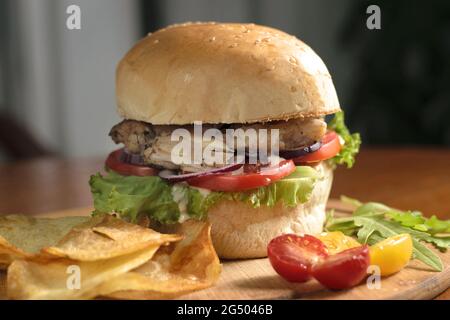 Großer Burger mit gebratenem Hühnerfleisch, Tomaten, Zwiebelringen und Salatblättern auf einem Holzbrett, mit Kartoffelchips, mit gelben und roten Kirschtomaten Stockfoto