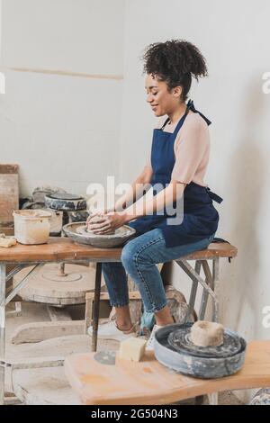 Lächelnde junge afroamerikanische Frau modelliert feuchten Ton auf dem Rad mit Händen in Keramik Stockfoto