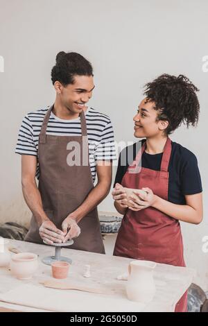 Lächelndes junges afroamerikanisches Paar in Schürzen, das Tontöpfe machte und sich in Töpferwaren ansah Stockfoto