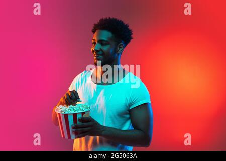 Bild eines jungen afrikanischen Mannes mit der Schachtel Popcorn in den Händen auf einem leuchtend rosa Neon-Hintergrund. Stockfoto