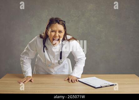 Wütend wütende Ärztin schreiend auf Kamera hinter Schreibtisch Studio Schuss stehen Stockfoto