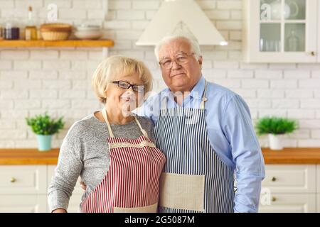 Porträt eines glücklichen verheirateten Seniorenpaares, das in Schürzen gekleidet und in der Küche posiert. Stockfoto