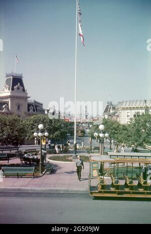 Disneyland, Kalifornien, 1959. Blick auf den Marktplatz von der Hauptstraße aus. Ein Fahnenmast fliegt die Stars & Stripes und die Flagge Kaliforniens, während sich die Besucher auf Bänken entspannen und vorbeikommen. Im Vordergrund wartet ein von Disneyland gezogener Straßenwagen, und in der Ferne sind die Main Street und das Dornröschenschloss zu sehen. Ein „Disneyland ‘59“-Banner wird auf der anderen Seite der Main Street angezeigt und wirbt für die Neuzugänge des Resorts für dieses Jahr. Stockfoto