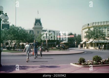 Disneyland, Kalifornien, 1959. Blick auf den Marktplatz und die Hauptstraße. Eine Familie überquert die Straße in Richtung Main Street Station, während im Hintergrund ein von Pferden gezogener Straßenwagen in Disneyland abgestellt wird. Ein Schild für den Disneyland Omnibus ist an einem Lampenpfosten angebracht. Die Wurlitzer Musikhalle ist an der Ecke Main Street und Town Square zu sehen. Ein „Disneyland ‘59“-Banner wird auf der anderen Seite der Main Street angezeigt und wirbt für die Neuzugänge des Resorts für dieses Jahr. Auf dem Platz ist eine antike Kanone zu sehen, während sich die Besucher auf Bänken entspannen und vorbeikommen. Stockfoto