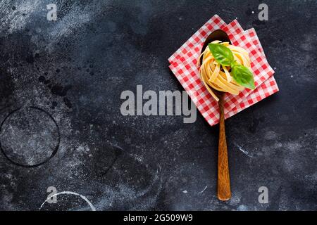 Frische hausgemachte Pasta-Tagliatelle auf Holzlöffel mit Pasta-Zutaten Tomaten, rohes Ei, Basilikumblatt auf dem dunklen Beton-Hintergrundtisch. Kochen c Stockfoto