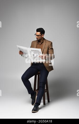 Muslimischer Geschäftsmann liest Geschäftszeitung auf Stuhl auf grauem Hintergrund Stockfoto