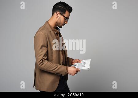 Seitenansicht eines jungen arabischen Geschäftsmannes, der Zeitung liest, mit isolierten Schriftzügen auf Grau Stockfoto
