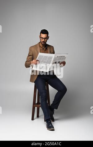 Arabischer Geschäftsmann liest Geschäftsnachrichten auf einem Stuhl auf grauem Hintergrund Stockfoto