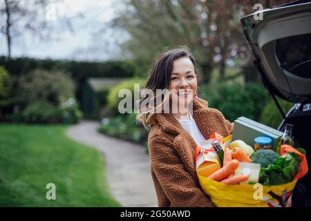 Eine junge Frau lächelt, blickt auf die Kamera und hält eine wiederverwendbare Tragetasche voller Lebensmittel. Sie entlädt das Auto nach dem Lebensmitteleinkauf. Stockfoto