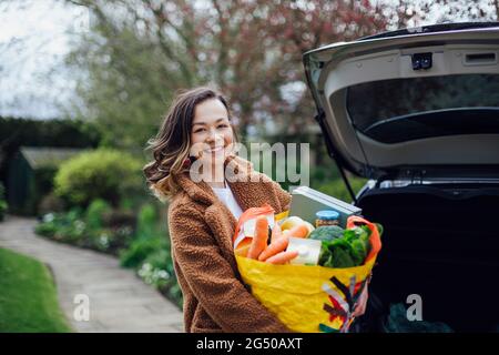 Eine junge Frau lächelt, blickt auf die Kamera und hält eine wiederverwendbare Tragetasche voller Lebensmittel. Sie entlädt das Auto nach dem Lebensmitteleinkauf. Stockfoto