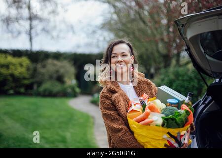 Eine junge Frau lächelt, schaut von der Kamera weg und hält eine wiederverwendbare Tragetasche voller Lebensmittel. Sie entlädt das Auto. Stockfoto